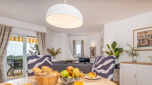 Un salon moderne avec un coin repas, comprenant une grande fenêtre, un canapé, une table à manger avec des fruits et du pain, et un luminaire suspendu.