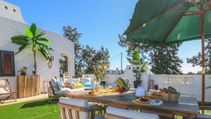 Un espace extérieur aménagé pour les repas, avec une table en bois, des chaises et un parasol vert. On y trouve également des plantes en pot et un mur blanc orné d’une fenêtre en arc de cercle.