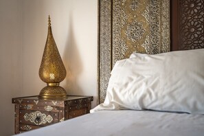 Un lit avec une literie blanche, une table de chevet en bois ornée de motifs complexes et une lampe traditionnelle en laiton.