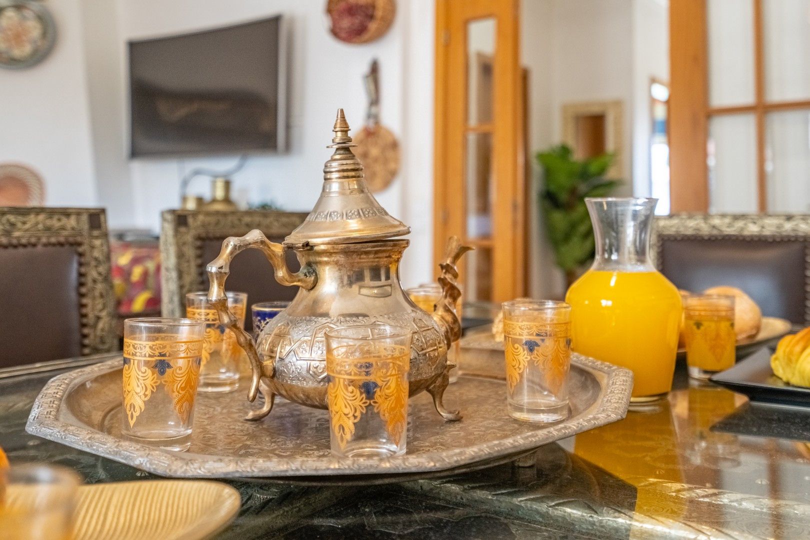A dining table with a traditional brass teapot, matching cups, and a pitcher of orange juice.