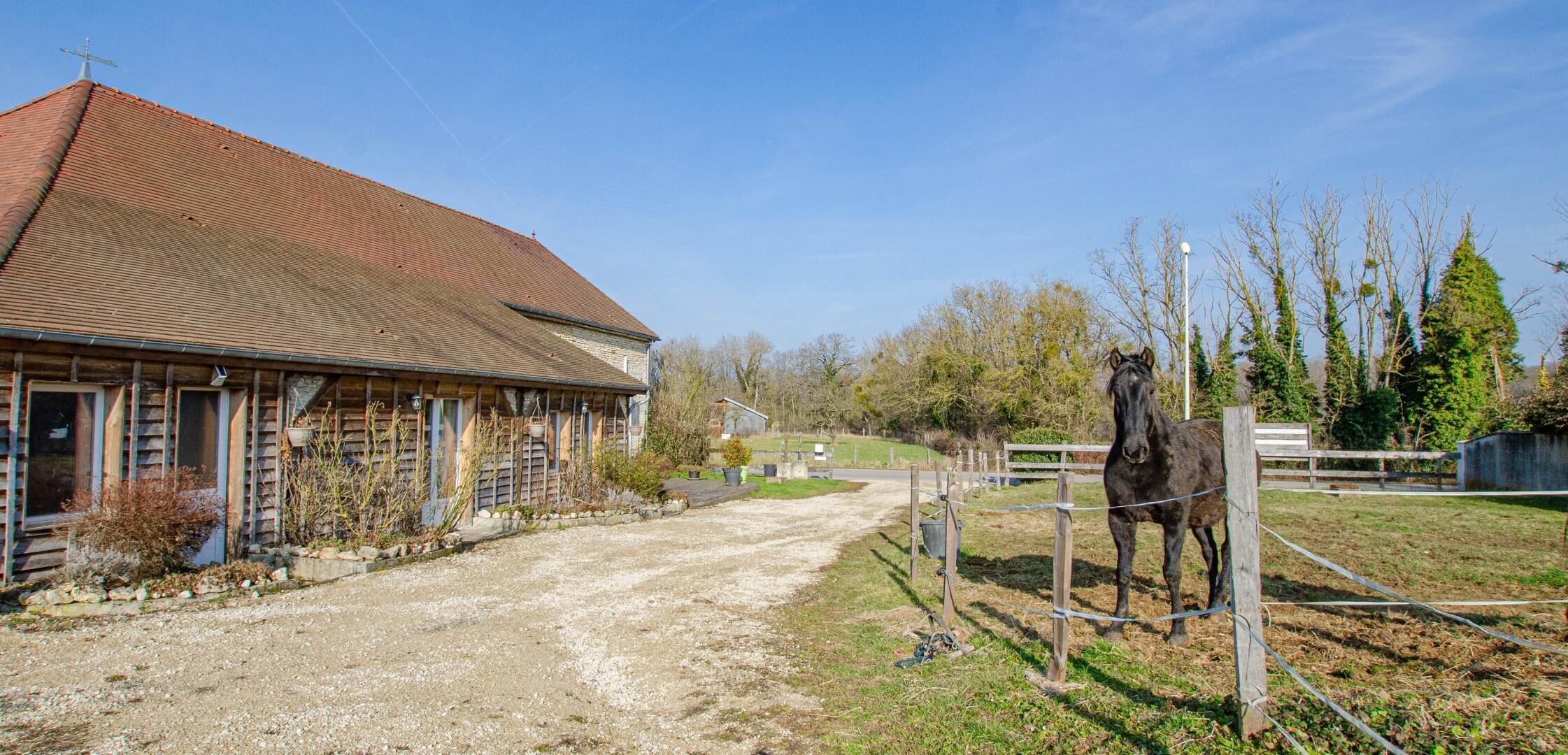 Une écurie avec un cheval, une allée en gravier et un bâtiment en bois avec un toit rouge.