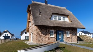 Ein traditionelles Haus mit Strohgedeck und einem hölzernen Boot davor, unter blauem Himmel.
