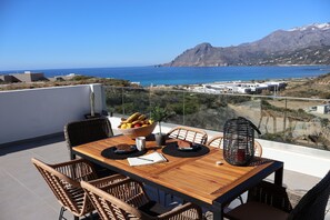 Una mesa de madera con sillas de mimbre en un balcón con vistas al mar y montañas a lo lejos.