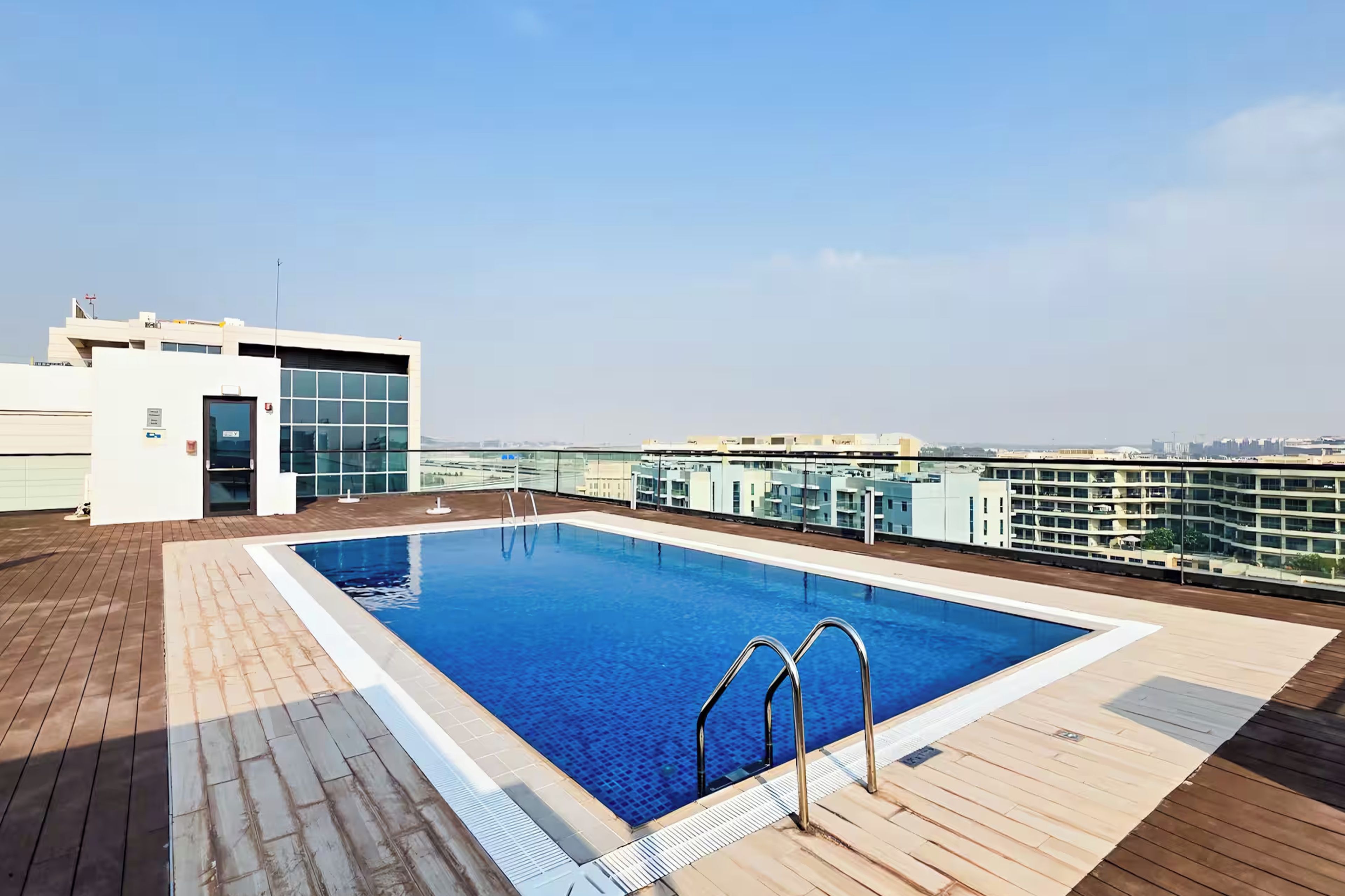A rooftop pool with clear blue water, surrounded by a wooden deck and a glass railing.