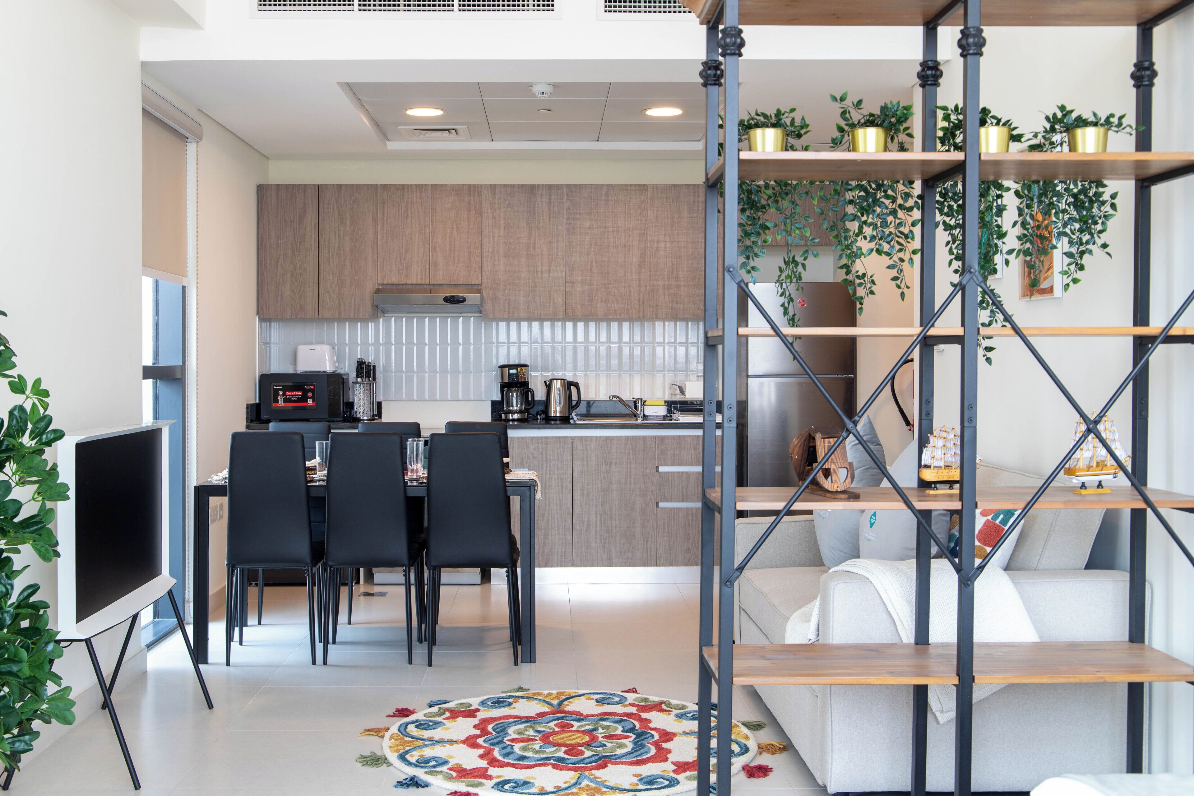Une cuisine moderne avec des meubles en bois, un coin repas avec des chaises noires, une machine à café et un téléviseur. On y trouve un escalier avec une rampe en métal et des étagères ornées de plantes.