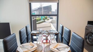 Un coin repas avec une table dressée pour quatre personnes, des chaises noires à revêtement en tissu et une vue sur un bâtiment en construction, visible à travers une fenêtre.