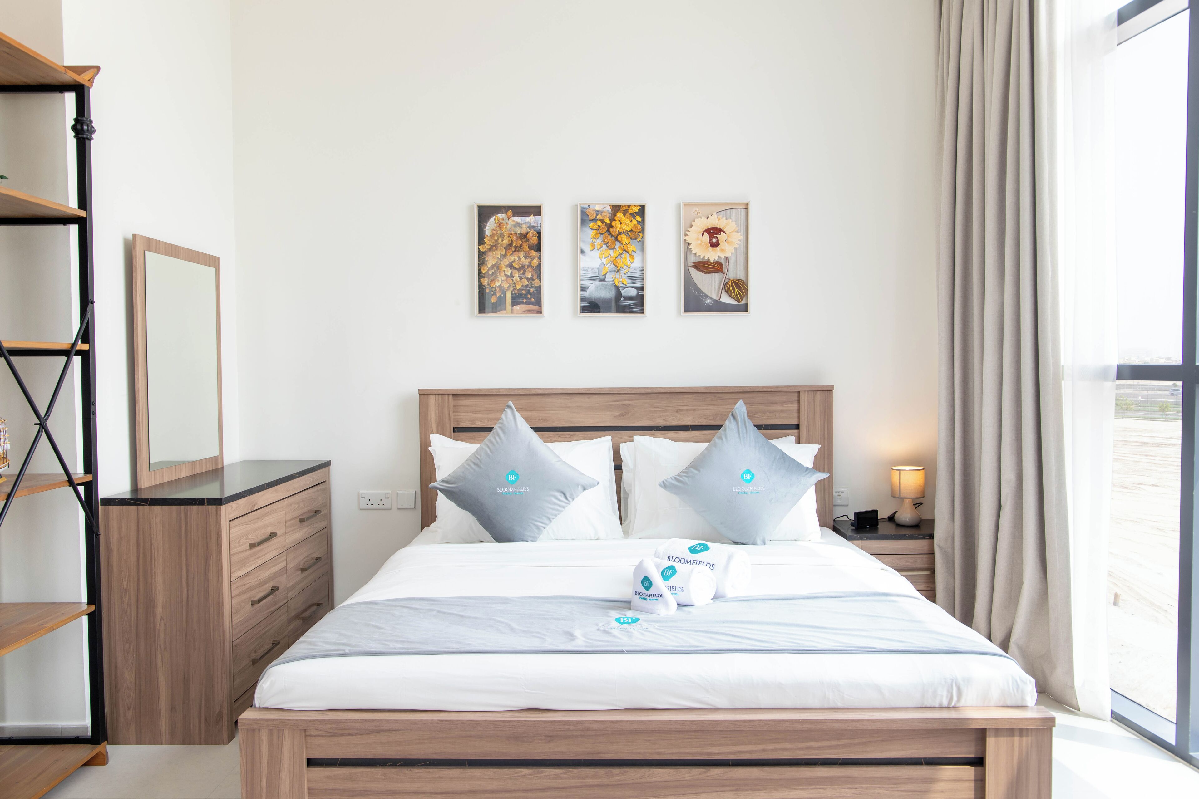 Een netjes ingerichte hotelkamer met een bed, nachtkastje, spiegel en een plank. Boven het bed hangen drie ingekaderde foto’s.