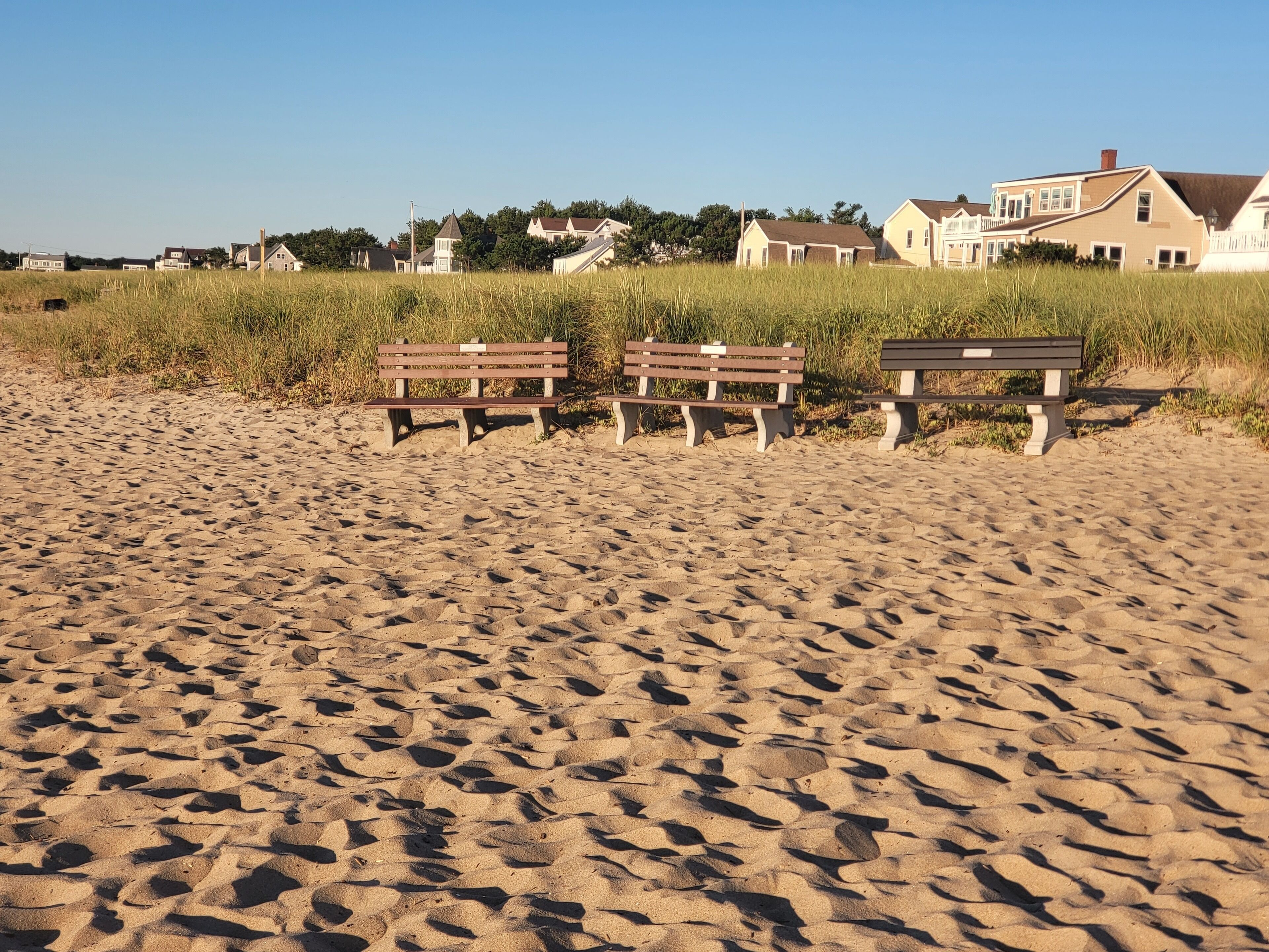 Een zandstrand met drie houten bankjes en huizen op de achtergrond.