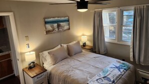 Een slaapkamer met een bed, twee nachtkastjes met lampen, een plafondventilator, een raam met gordijnen en een schilderij aan de muur.