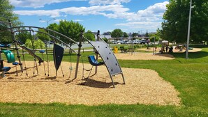 A playground with swings, slides, and climbing structures on a grassy area with a parking lot in the background. - Splendid, two-bedroom, one-bathroom condo,  two short blocks from the beach! (Old Orchard Beach)