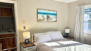 Une chambre à coucher avec un lit, deux tables de chevet avec des lampes, un tableau représentant une scène de plage, une armoire en bois et une fenêtre avec des rideaux.