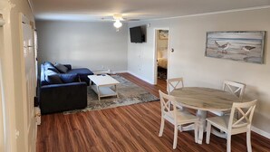 A living room with a dark blue sofa, a white coffee table, and a wooden dining table with four chairs. There is a painting of birds on the wall and a television mounted on the wall. - Stunning, two-bedroom, one-bathroom condo,  two short blocks from the beach! (Old Orchard Beach)