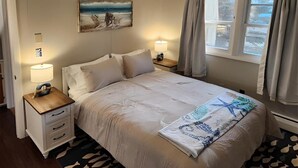 A neatly arranged bedroom with a bed, bedside tables, a lamp, a painting, and a window with curtains. - Stunning, two-bedroom, one-bathroom condo,  two short blocks from the beach! (Old Orchard Beach)