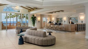 Un vaste espace de vie comprenant un grand canapé, un coin repas avec des chaises, une cuisine avec un bar et un balcon offrant une vue sur les palmiers et l’océan.