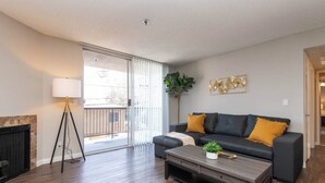 Un salon moderne avec un canapé de couleur sombre, une table basse et une lampe. On trouve une cheminée sur la gauche et une grande fenêtre avec des stores.