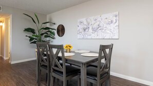 Un coin repas avec une table dressée pour quatre personnes, entourée de chaises en bois. On peut voir une grande plante en pot et une œuvre d’art accrochée au mur.