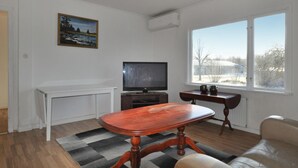 Een woonkamer met een televisie, een schilderij, een raam met uitzicht en een houten tafel.
