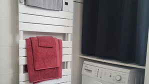 Uma área de lavanderia com um varal branco fixado na parede, contendo duas toalhas, uma máquina de lavar frontal e uma cortina preta cobrindo parcialmente uma janela.