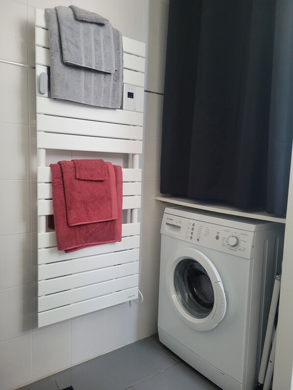 A laundry area with a white wall-mounted drying rack holding two towels, a front-loading washing machine, and a black curtain partially covering a window. - Self-catering cottage, air-conditioned, between the Loire Valley and Châteaux (Blois)