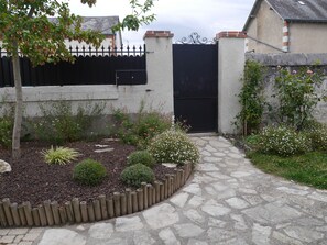 Ein Steinweg führt zu einem schwarzen Tor, umgeben von einem Garten mit verschiedenen Pflanzen und einem Holzzaun.