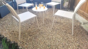 Um pátio externo com quatro cadeiras brancas e uma pequena mesa redonda, cercado por cascalho.