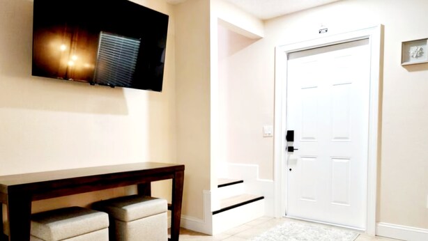 A hallway with a white door, a mounted television, a dark wooden bench with storage, and a light-colored rug. - Toria Suites 920 (Lakeland)