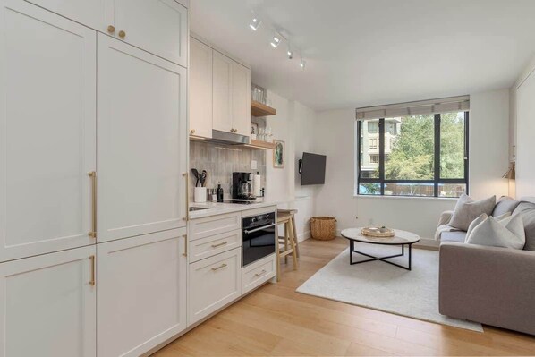 A modern kitchen with white cabinetry, a coffee machine, and a dining area with a grey sofa and a coffee table.