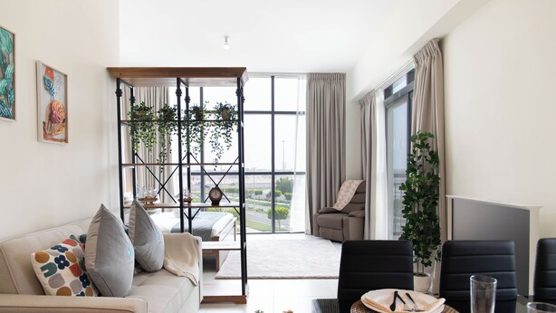 A modern living room with a sofa, a dining table set for two, and a large window offering a view of the outdoors. - Bloomfields Artistic STU in The View (Abu Dhabi)