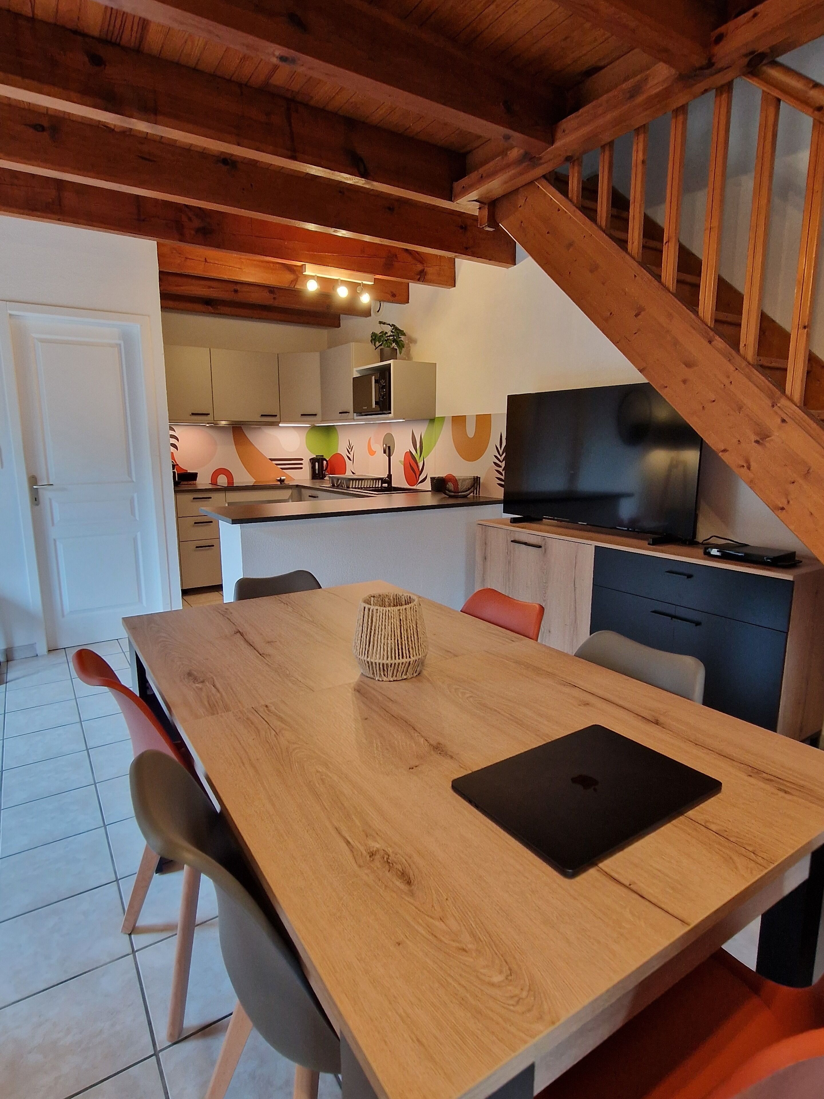 A dining area with a wooden table, chairs, and a laptop. A staircase leads to a kitchen with a TV and cabinets.