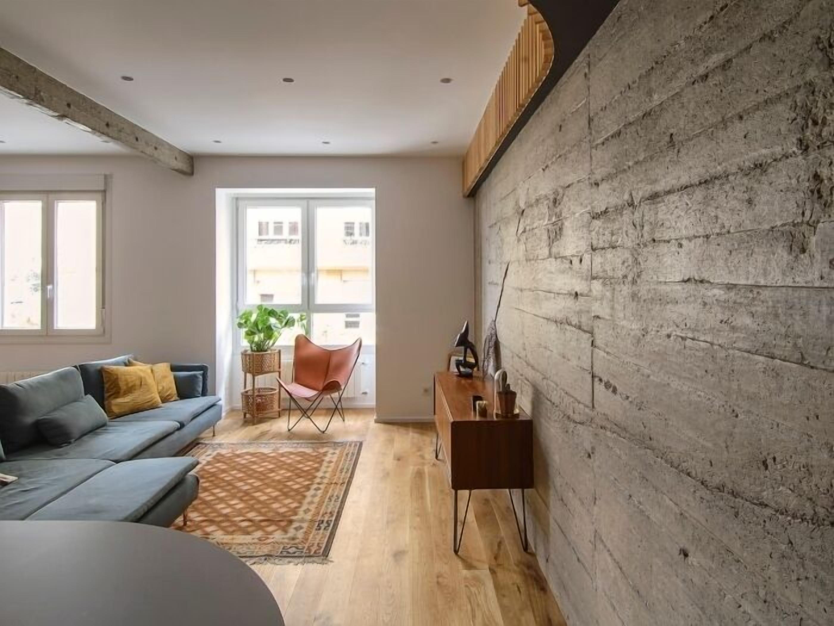 Un salon moderne avec un canapé gris, une table basse en bois et une grande fenêtre donnant sur des immeubles.
