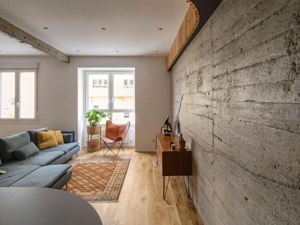 A modern living room with a grey sofa, a wooden coffee table, and a large window with a view of buildings.