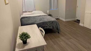 Una habitación pequeña y ordenada con una cama, una mesa con una planta en maceta y una puerta.
