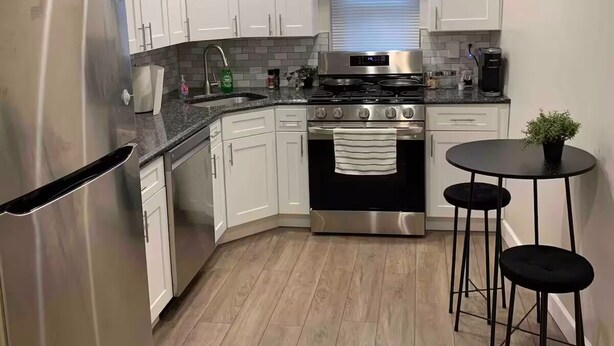 Una cocina moderna con armarios blancos, electrodomésticos de acero inoxidable, una encimera negra y una pequeña mesa redonda con taburetes.