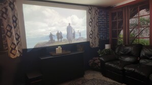 Un saló amb un televisor gran que mostra una escena natural, un sofà de cuir negre, una taula petita amb espelmes i una finestra amb cortines estampades.