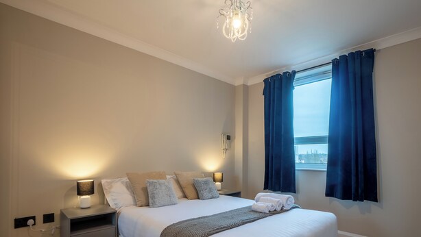 A neatly arranged bedroom with a bed, bedside tables, a lamp, and a view through a window with curtains.