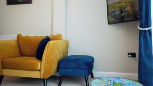 A living room with a yellow sofa, a blue ottoman, a round table with a colorful pattern, and two framed pictures on the wall.
