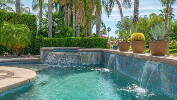 A pool with a waterfall feature, surrounded by palm trees and various potted plants. - Two Resort Style Homes Steps from Each Other, 7 Bedrooms, Pool, Hot Tub, Misters (Cathedral city)