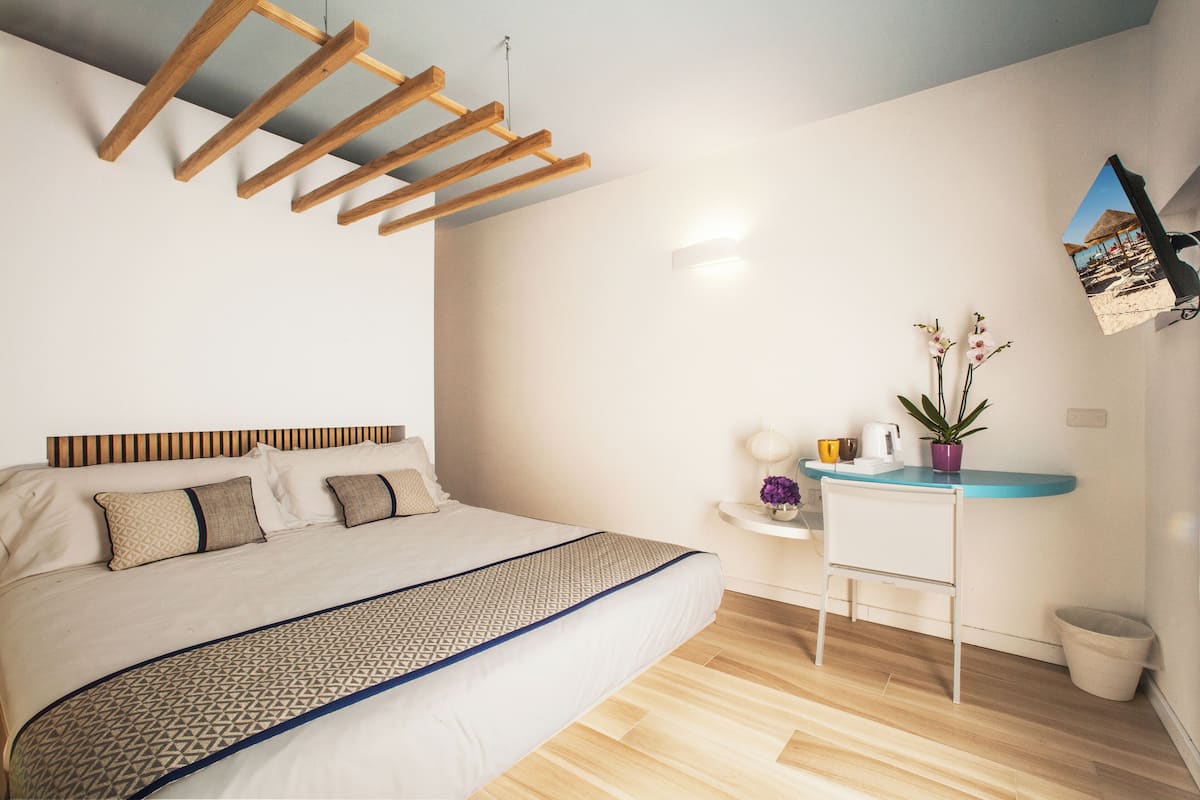 A modern bedroom with a wooden ceiling, a bed with patterned bedding, a small desk with a plant, and a wall-mounted picture.