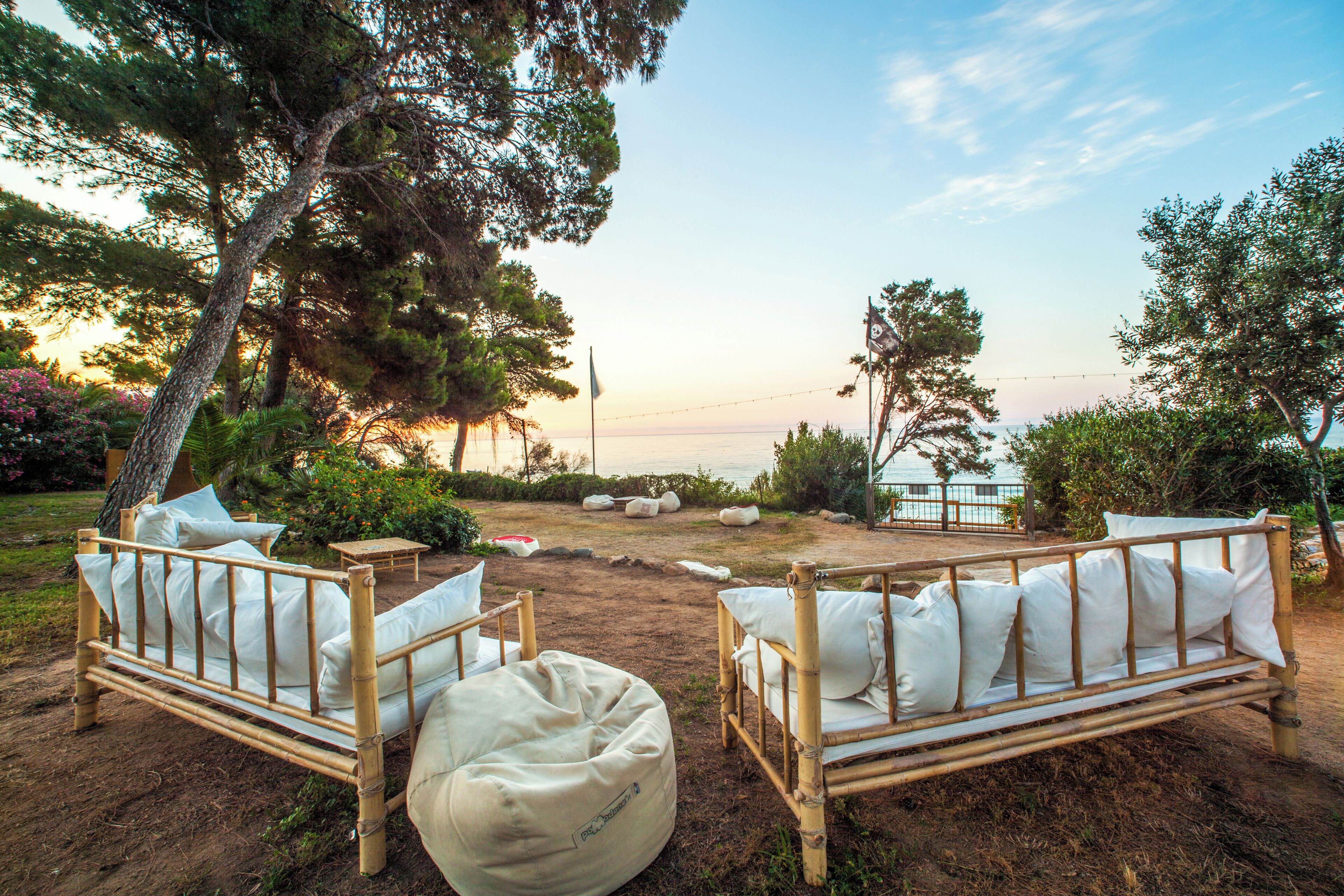 Dos camas de día de madera con cojines blancos, ubicadas en una zona arenosa con vista al mar y árboles al fondo.