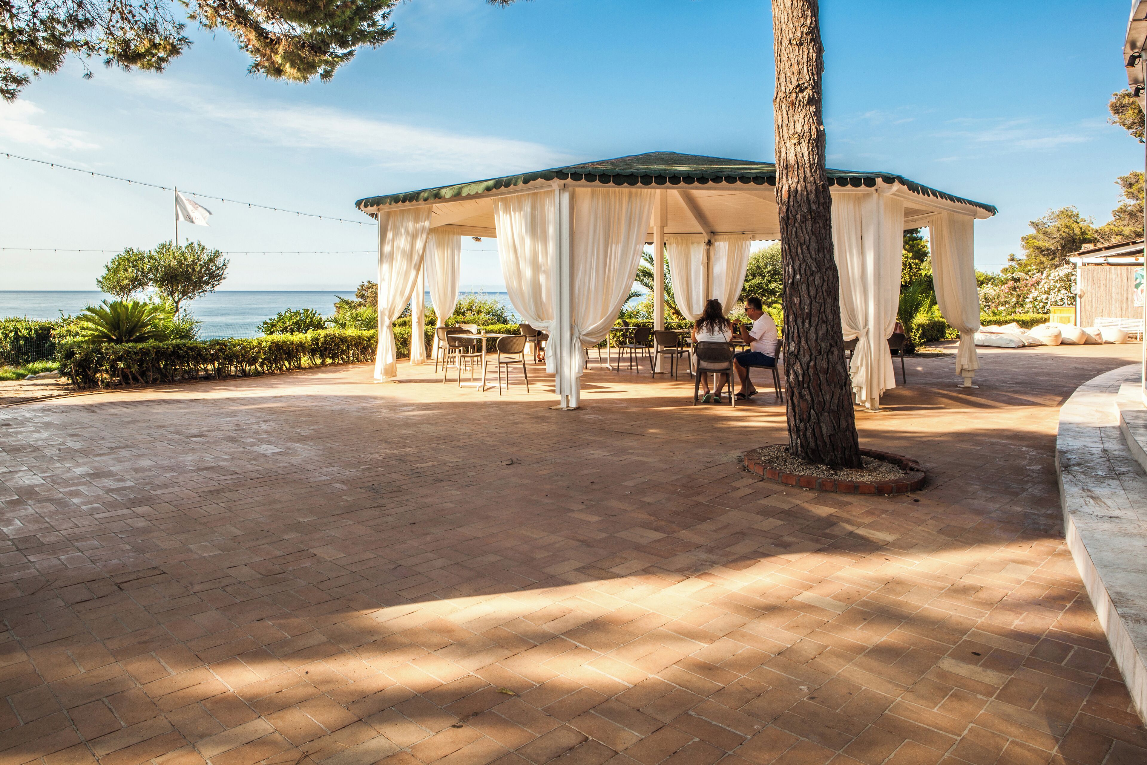 A covered outdoor seating area with a view of the sea, featuring a gazebo with white curtains and brick flooring.
