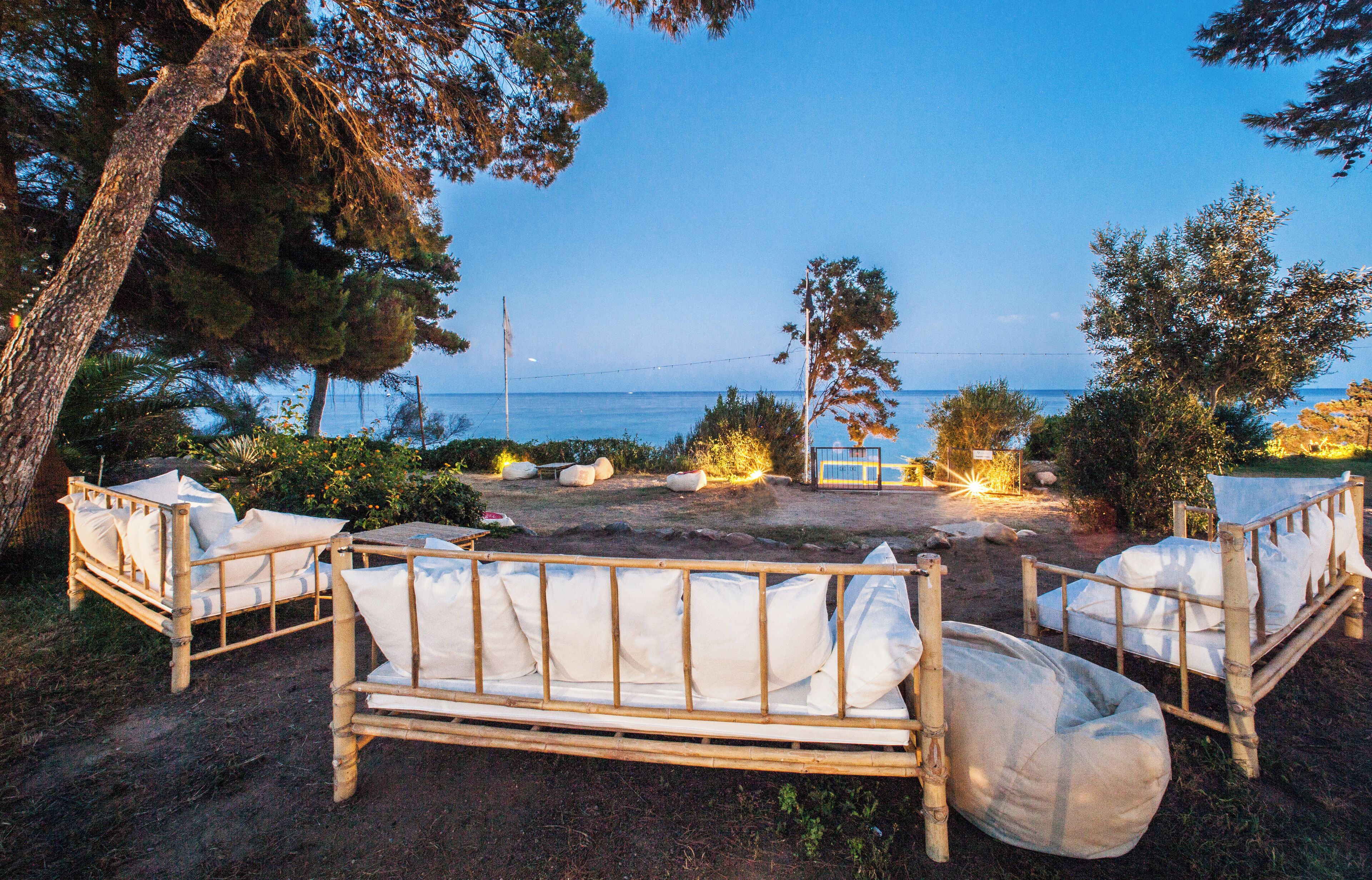 A wooden bench with cushions and a small table, set against a backdrop of trees and a view of the sea.