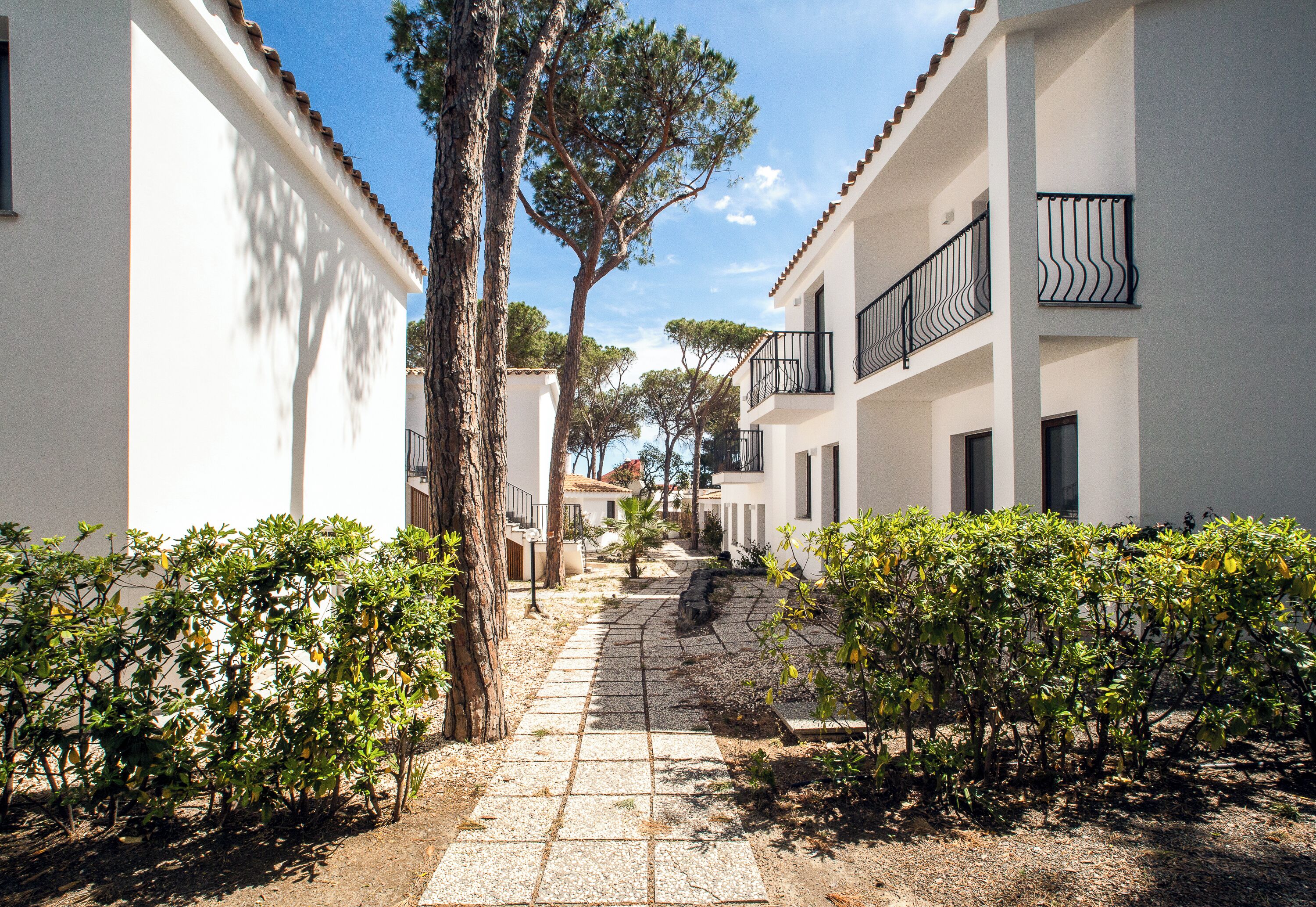 A narrow pathway flanked by white buildings with balconies, leading to a tree-lined area.