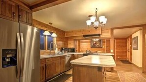 A modern kitchen with wooden cabinets, a stainless steel refrigerator, a large island, and a chandelier.
