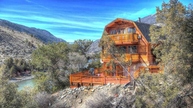 A two-story log cabin with a balcony, surrounded by a mountainous landscape and a river.