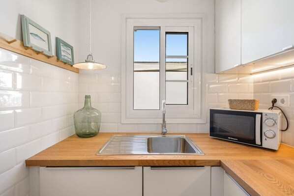 Una cocina moderna con armarios blancos, encimera de madera, pileta de acero inoxidable, microondas y lámpara colgante.