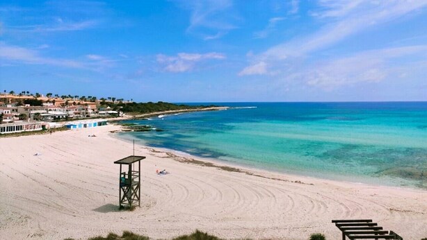 A beach with clear turquoise water, a lifeguard tower, and buildings in the background. - Villa Fontana, little paradise (Punta Prima)