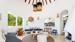 A modern living room with a sofa, coffee table, and ottoman, a kitchen area with bar stools, and a fireplace. - Villa Fontana, little paradise (Punta Prima)