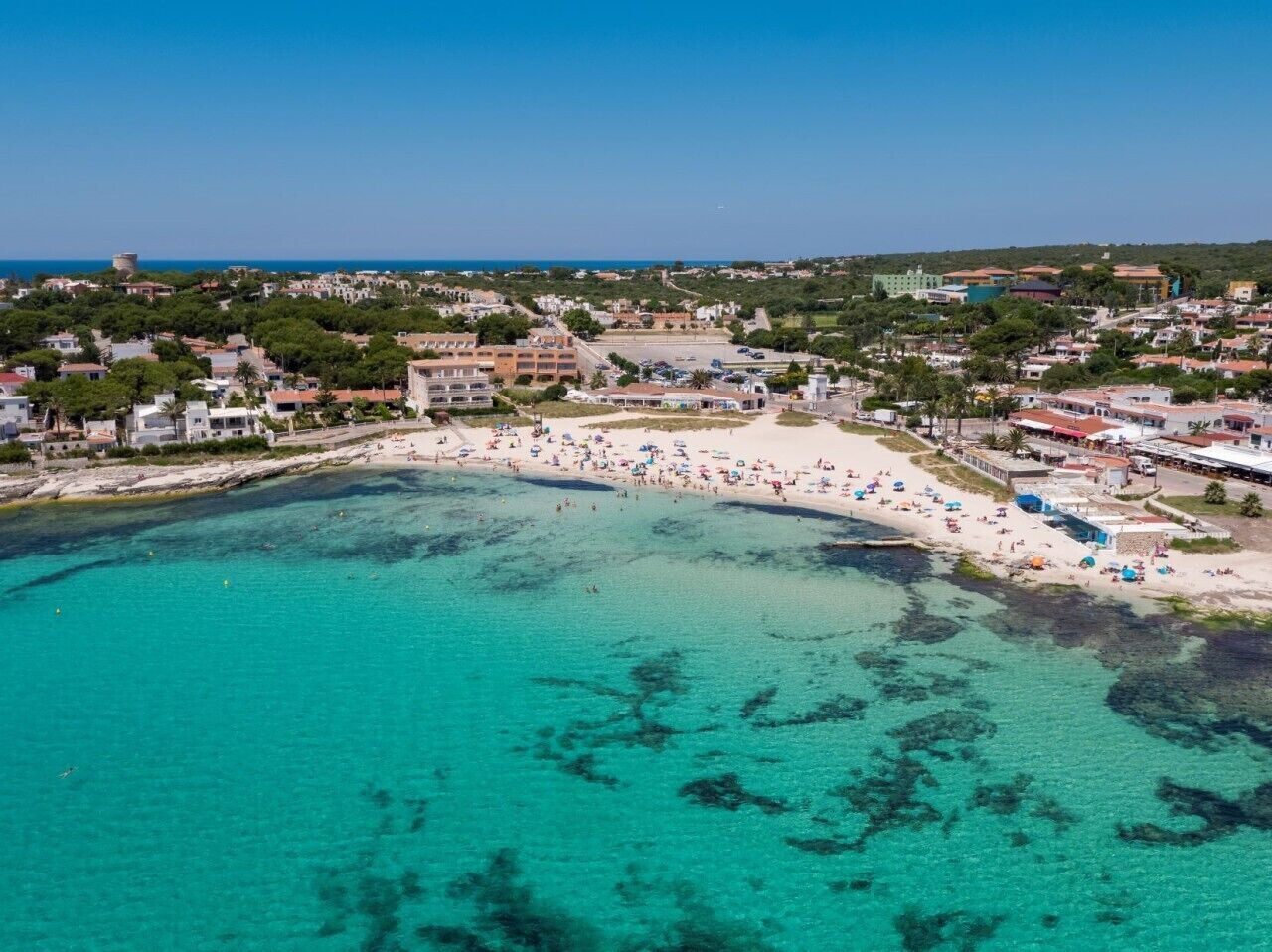 Vista aérea de un pueblo costero con una playa, aguas turquesas cristalinas y edificios al fondo.