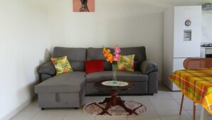 A living room with a grey sectional sofa, a small round table with a vase of flowers, and a dining area with a table covered by a yellow and red checkered tablecloth.