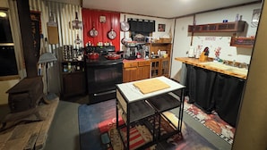 Eine kompakte Küche mit Herd, Backofen und verschiedenen Geräten. Ein kleiner Esstisch mit Schneidebrett ist vorhanden. Die Küche verfügt über Holzschränke und eine geflieste Spritzschutzwand.